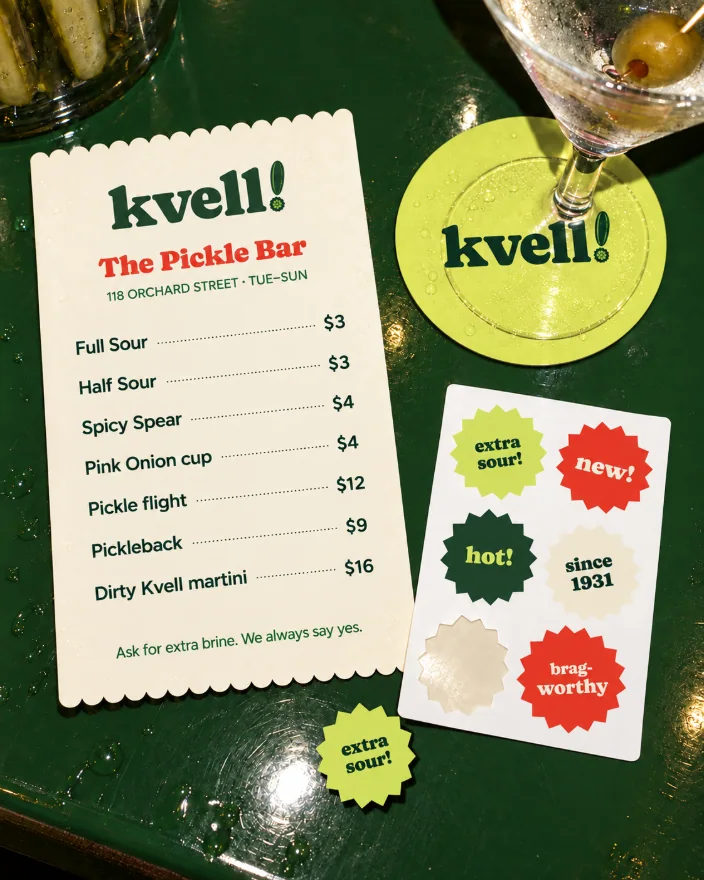 The pickle bar’s printed pieces on a deep green counter: a crinkle-edged menu on deli paper, a chartreuse coaster under a martini, and a sheet of starburst stickers with one peeled off onto the counter
